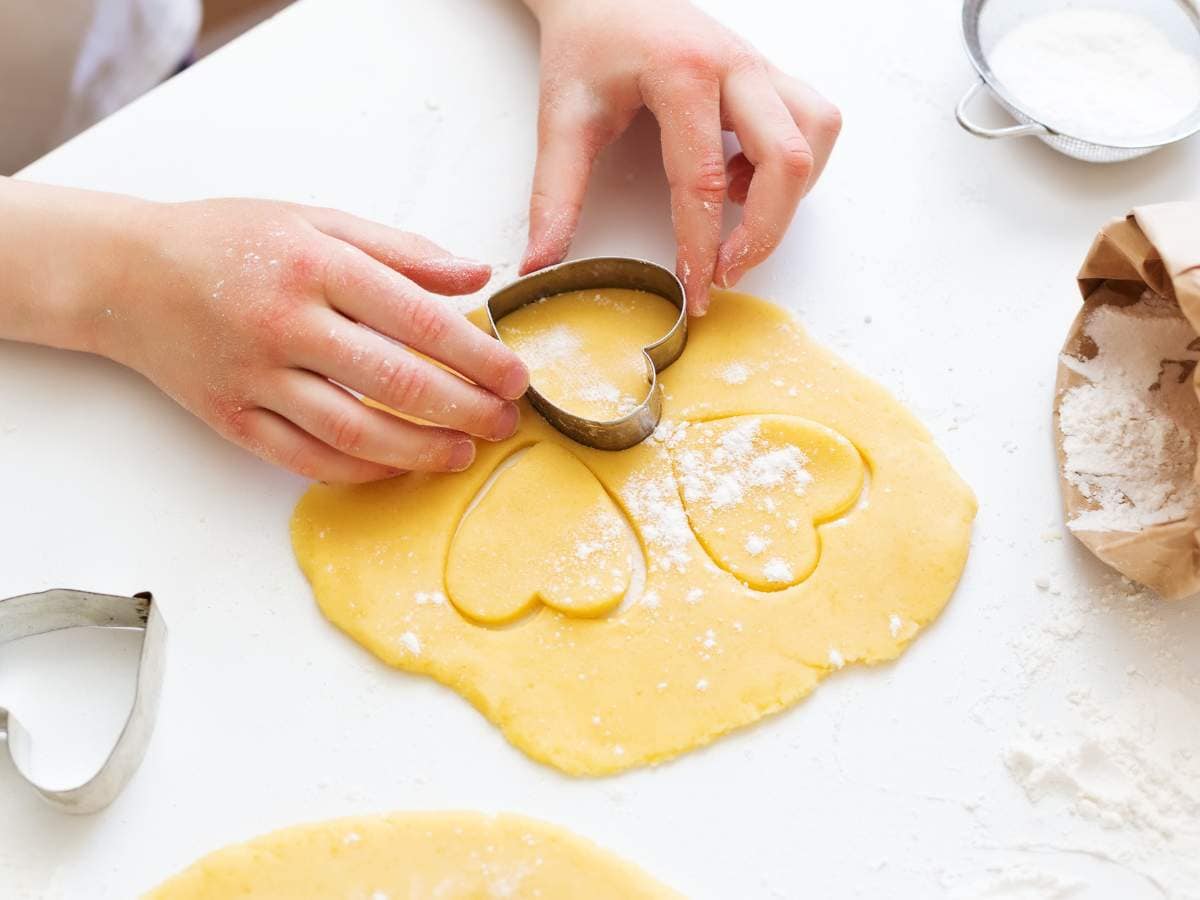 Hands pressing a heart-shaped cookie cutter into rolled dough, with flour scattered around.