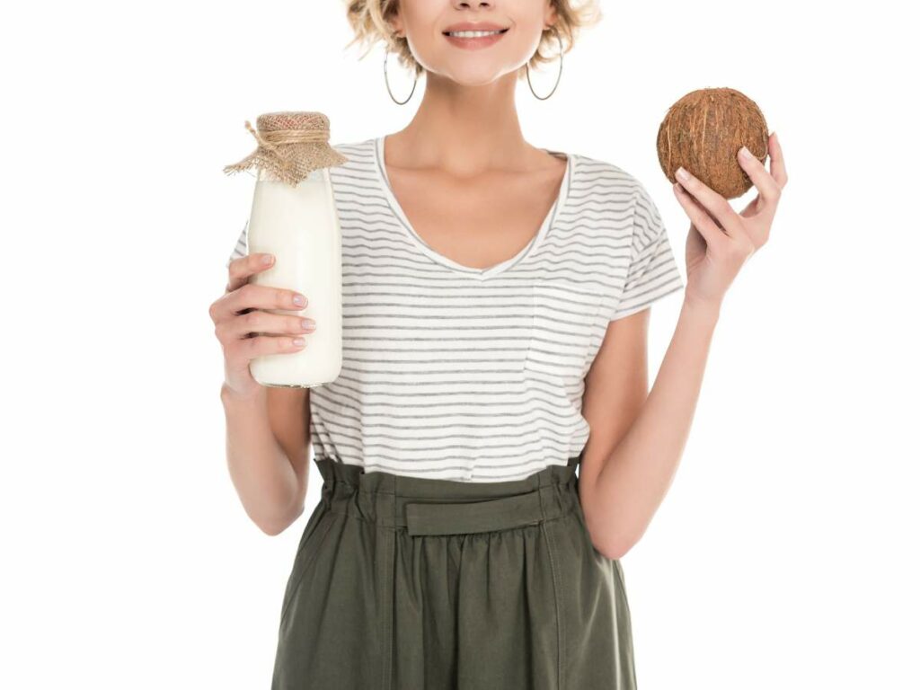Woman smiling, holding a bottle of milk and a coconut, dressed in a striped t-shirt and green skirt, isolated on a white background.