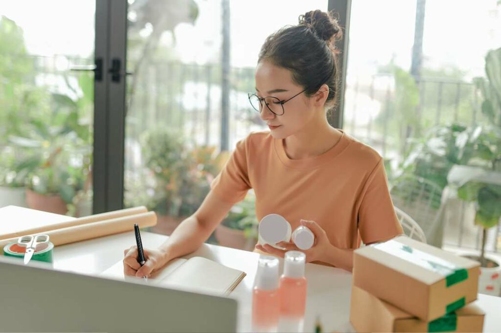 A woman wearing glasses writing in a notebook at a desk with shipping materials and products nearby.