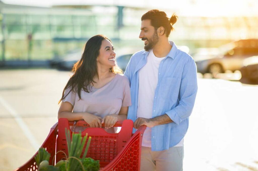 A couple smiling at each other while pushing a shopping cart in a parking lot.