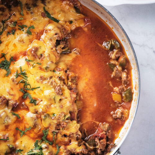 A skillet of cheesy meat with bell peppers, garnished with herbs on a marble surface.