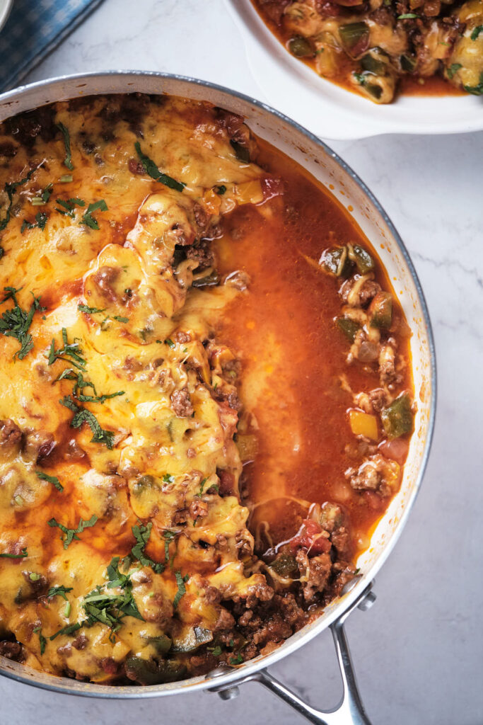 A skillet of cheesy meat with bell peppers, garnished with herbs on a marble surface.