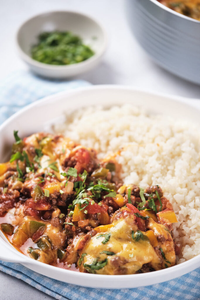 A plate of spicy minced meat with mixed vegetables and melted cheese, served alongside cauliflower rice, garnished with fresh herbs.