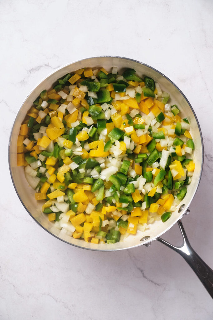 A stainless steel skillet on a white surface containing diced green bell peppers, yellow bell peppers, and onions.