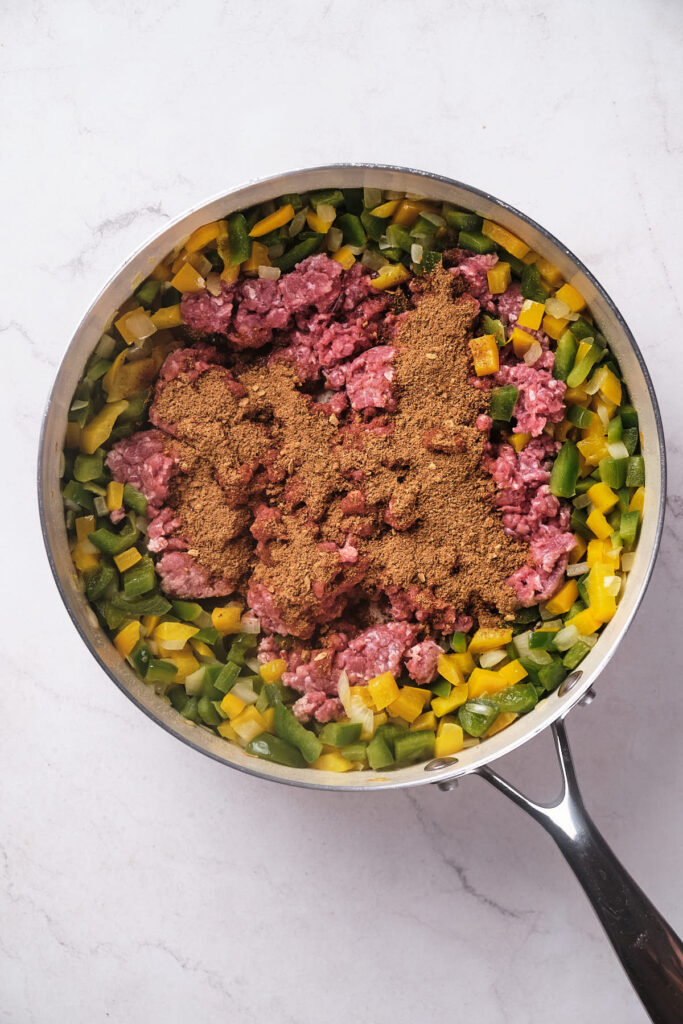 A skillet containing ground beef, green bell peppers, and yellow peppers, topped with a mound of taco seasoning.