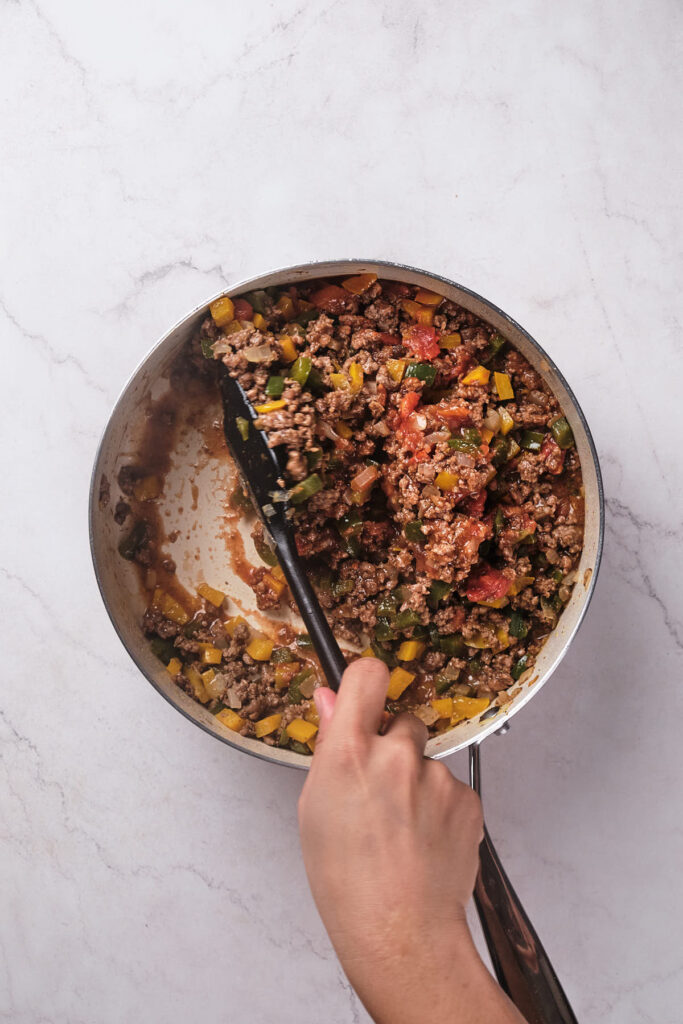A person's hand stirring a mix of ground meat, peppers, and vegetables in a skillet on a marble countertop.