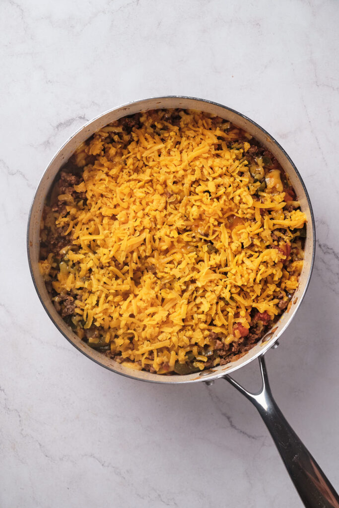 A skillet filled with seasoned ground beef and vegetables, topped with a layer of shredded cheddar cheese.