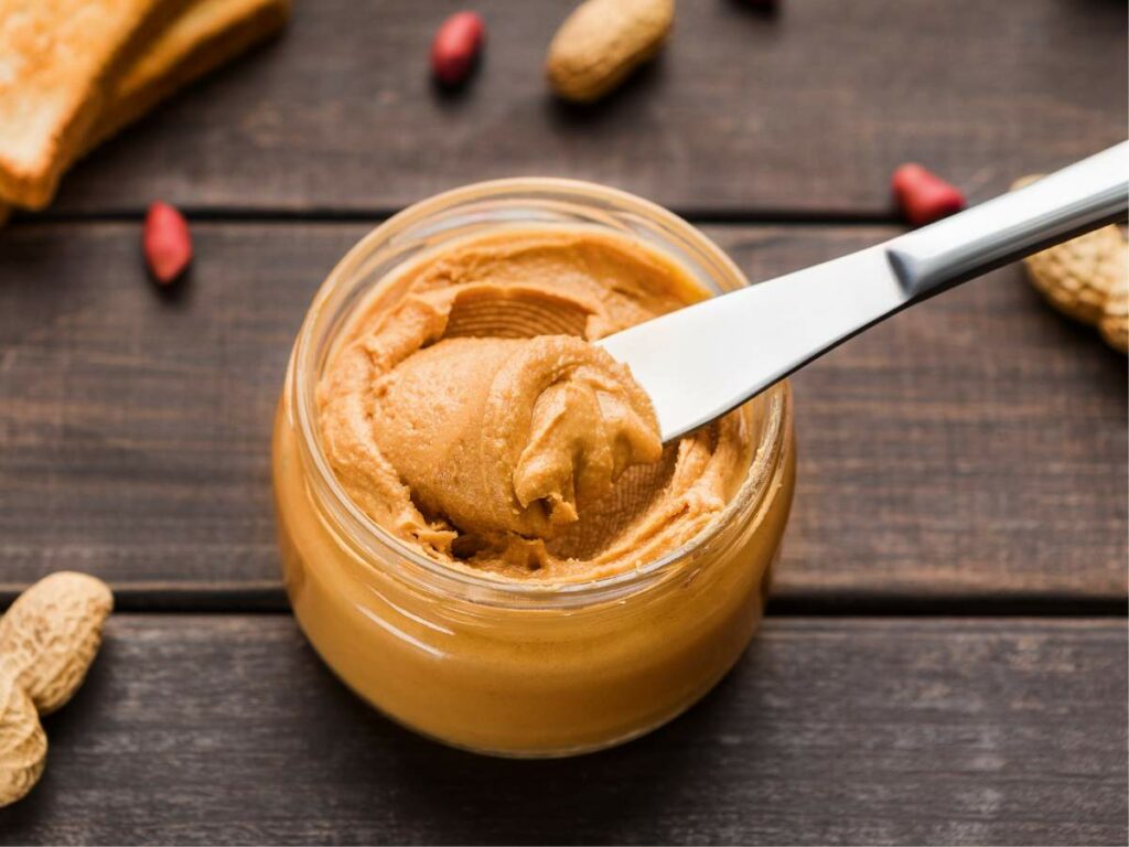 A knife spreading peanut butter from a glass jar, with whole peanuts and a loaf of bread on the side.