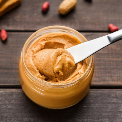 A knife spreading peanut butter from a glass jar, with whole peanuts and a loaf of bread on the side.