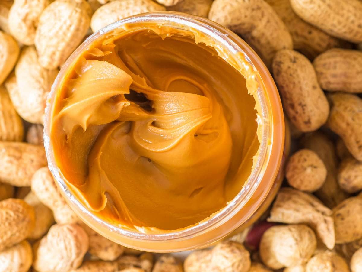 An open jar of creamy peanut butter surrounded by whole peanuts in their shells.