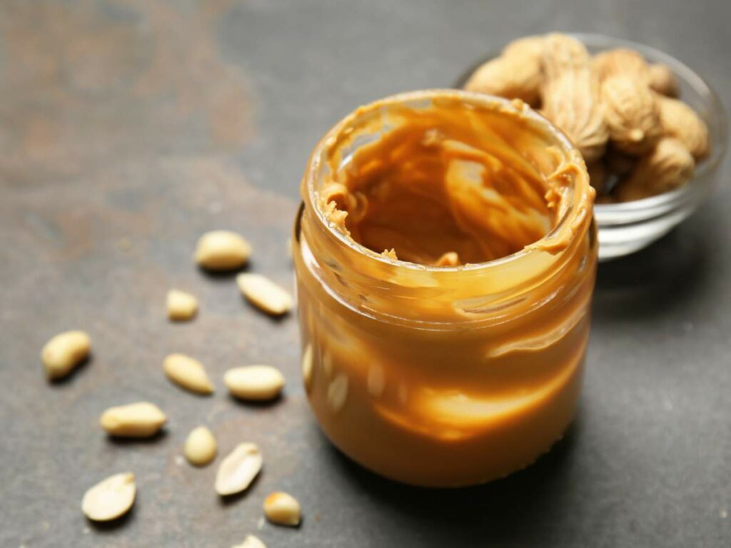 Open jar of peanut butter with peanuts scattered around on a dark surface.