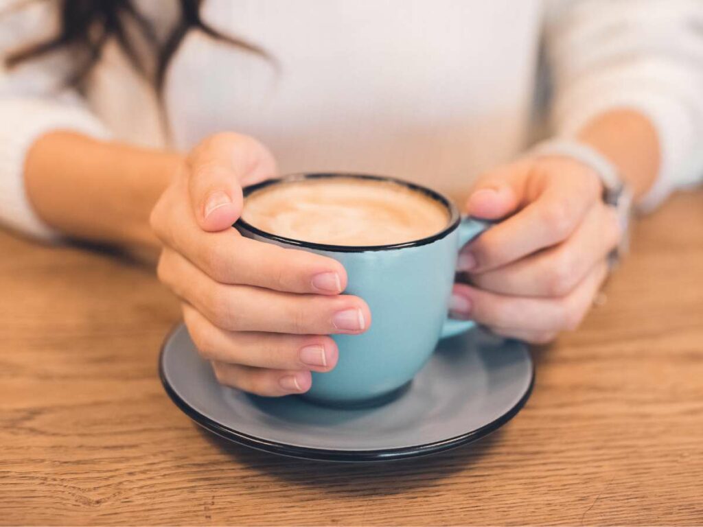 A woman holds a blue coffee cup with both hands, sitting at a wooden table.