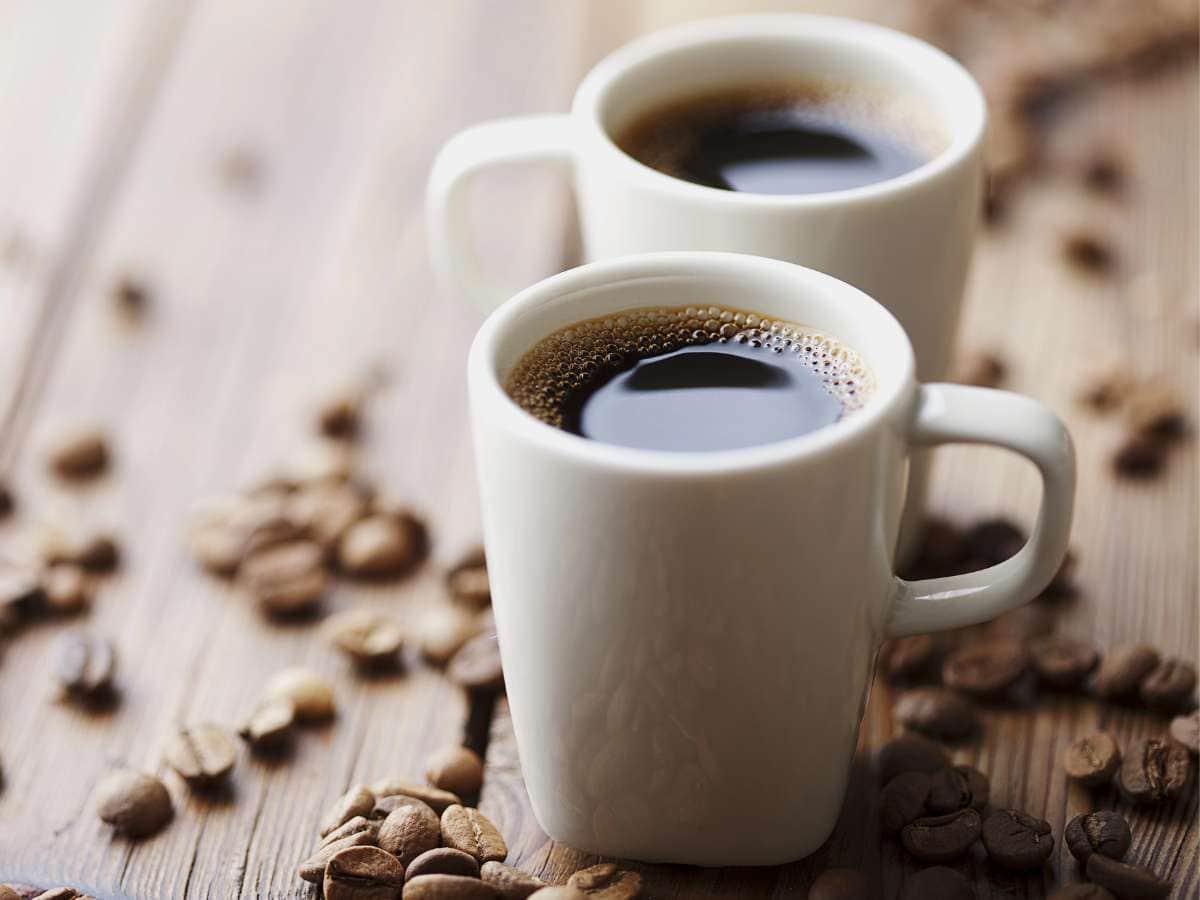 Two white cups filled with coffee surrounded by coffee beans on a wooden surface.