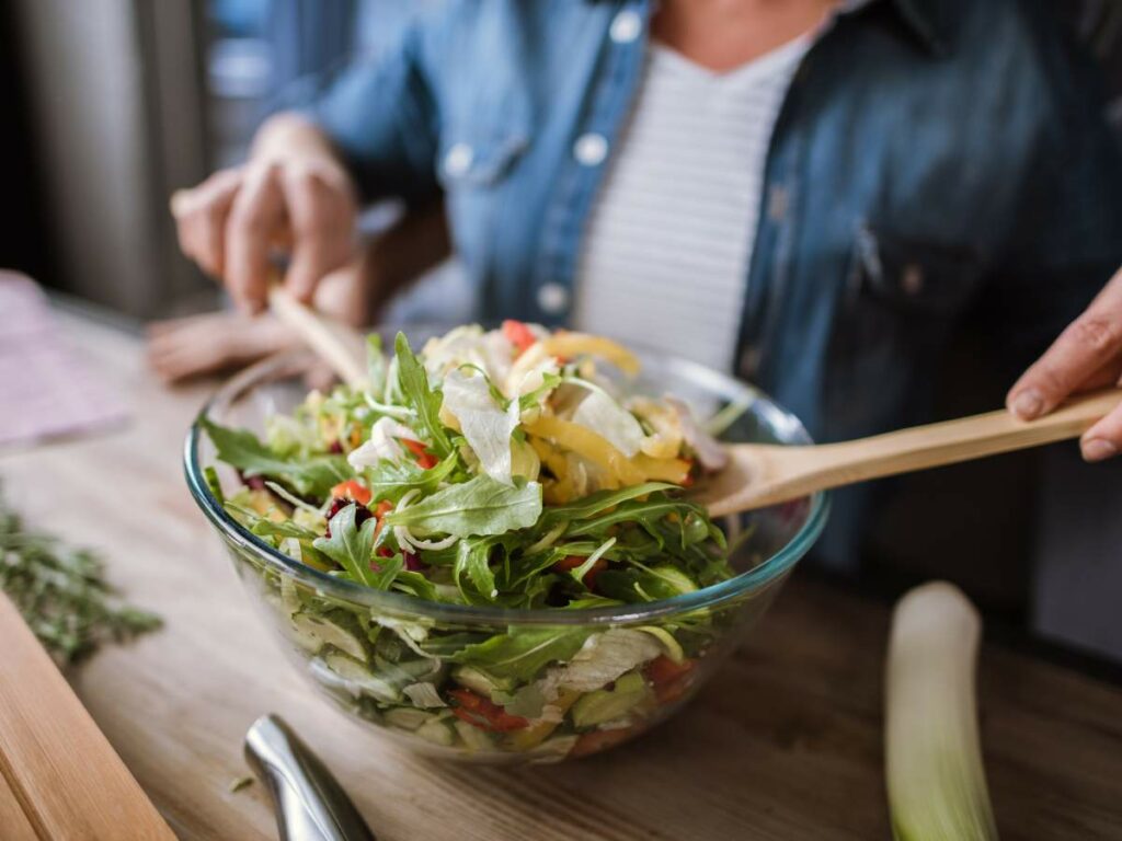Person serving fresh mixed salad from a glass bowl using wooden salad tongs at a wooden table.