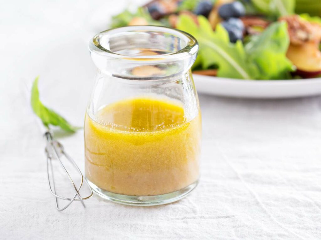 A small glass jar of homemade vinaigrette dressing with a metal whisk, beside a plate of salad with greens and grilled peaches.