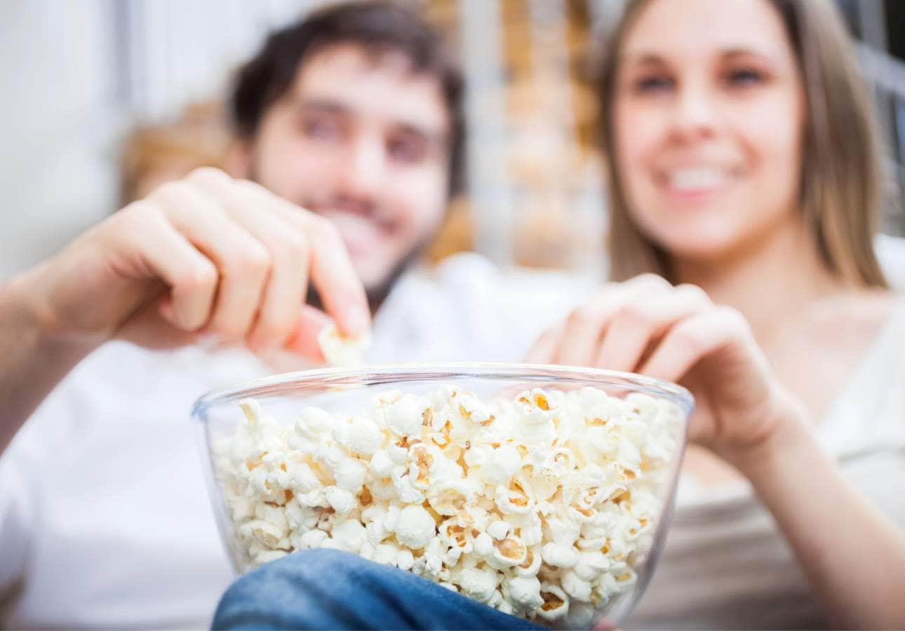 A couple sharing a bowl of popcorn, smiling and sitting closely together on a couch, with a focus on the popcorn bowl.