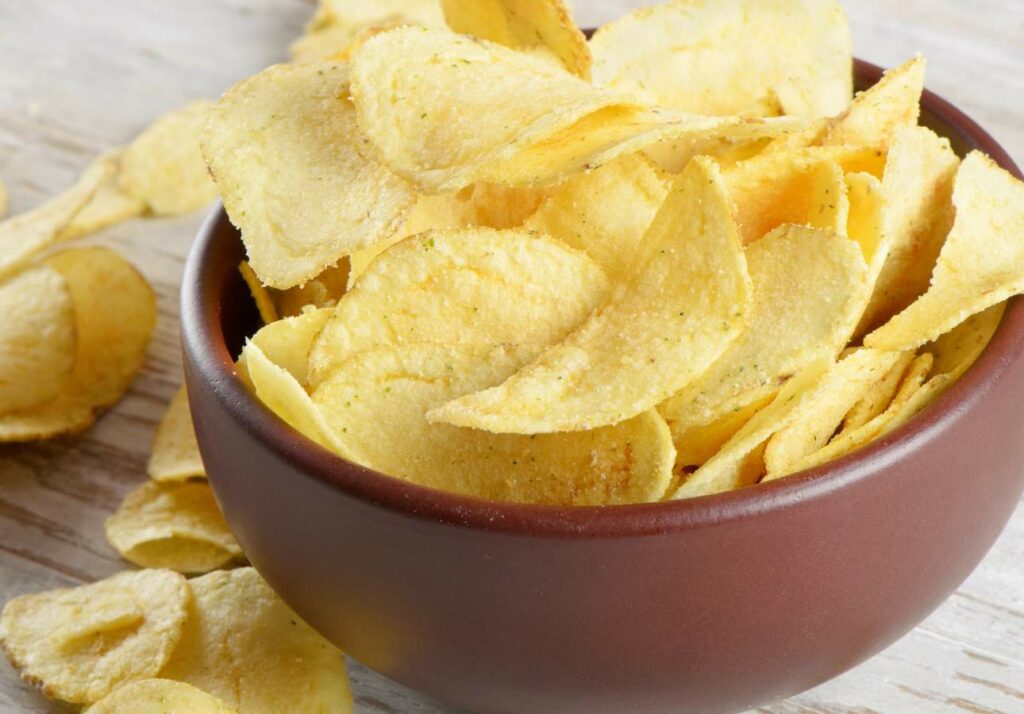 A bowl of crispy golden potato chips on a wooden surface, with some chips scattered around the bowl.