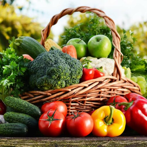A wicker basket filled with fresh vegetables and fruits including tomatoes, broccoli, cucumbers, and apples.