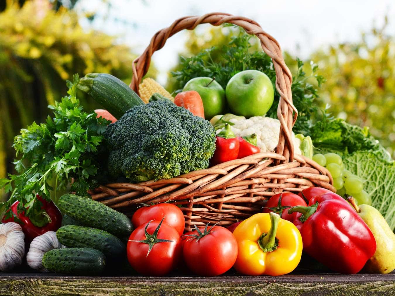 A wicker basket filled with fresh vegetables and fruits including tomatoes, broccoli, cucumbers, and apples.