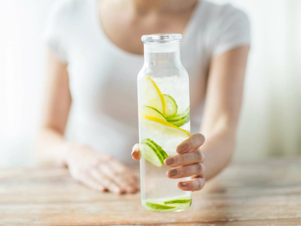 A person holding a clear bottle of water infused with lemon and cucumber slices, standing behind a wooden table.