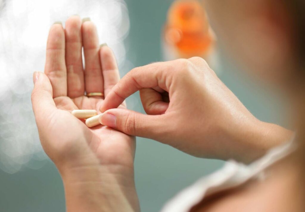 Woman holding two capsules in her hand with a blurred orange pill bottle in the background.