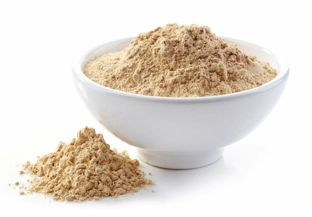 A white bowl filled with beige powder, with a small pile spilled beside it on a white background.