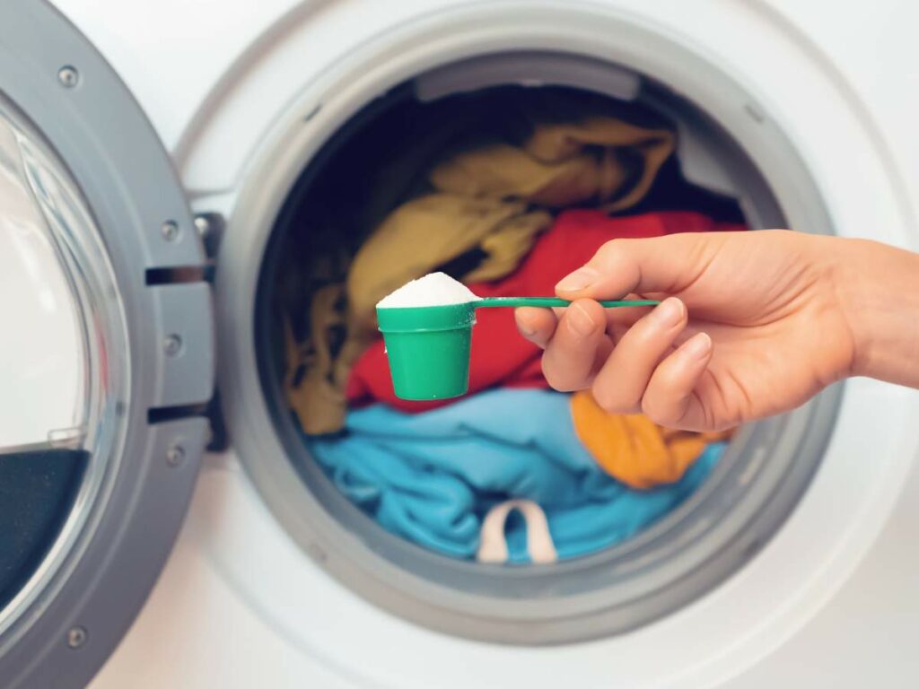 Hand holding a measuring scoop with detergent over an open washing machine filled with colorful clothes.