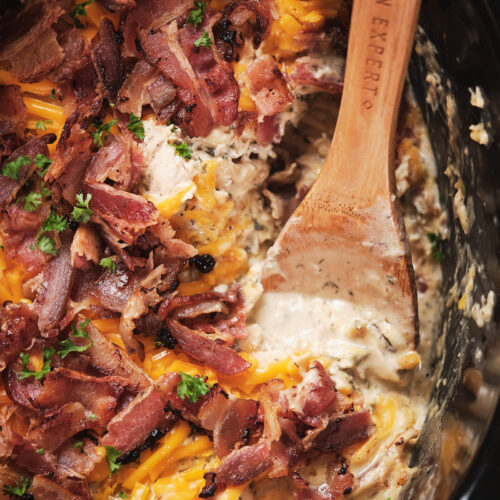 Close-up of a creamy chicken and cheese dish topped with crispy bacon and parsley in a slow cooker, with a wooden spoon.