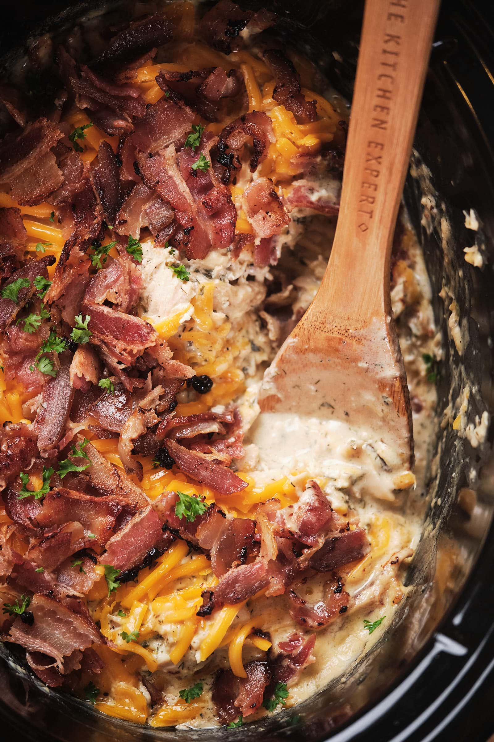 Close-up of a creamy chicken and cheese dish topped with crispy bacon and parsley in a slow cooker, with a wooden spoon.