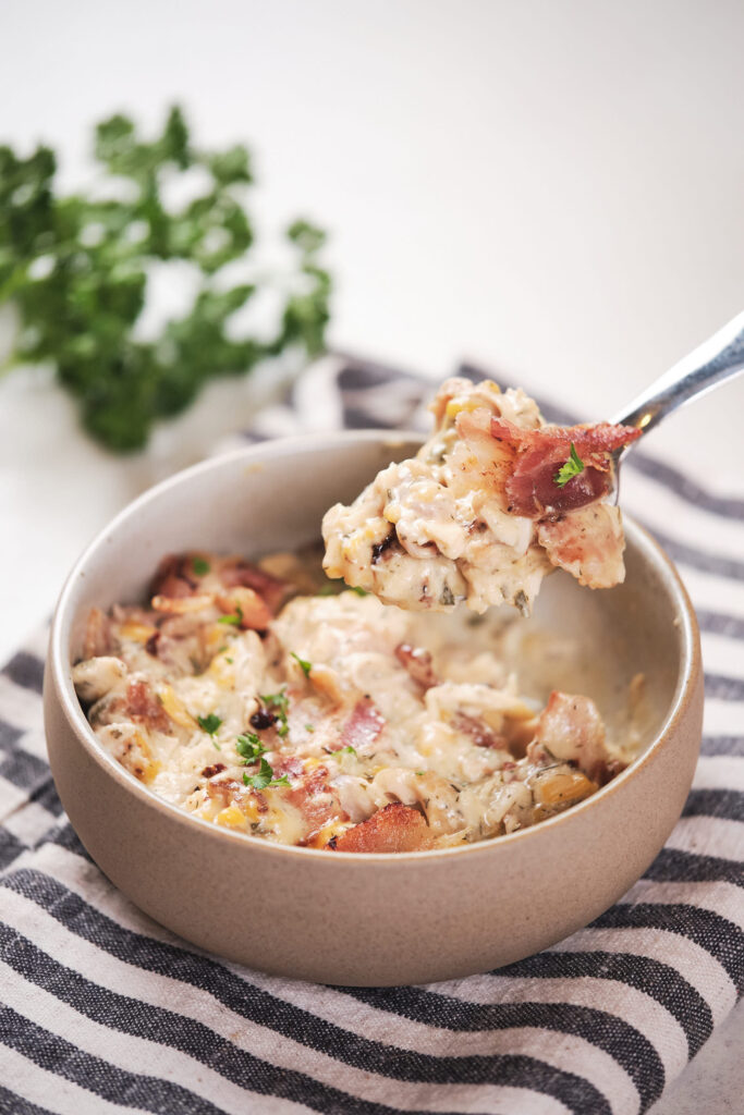 A spoon lifting a serving of creamy crack chicken from a beige bowl.