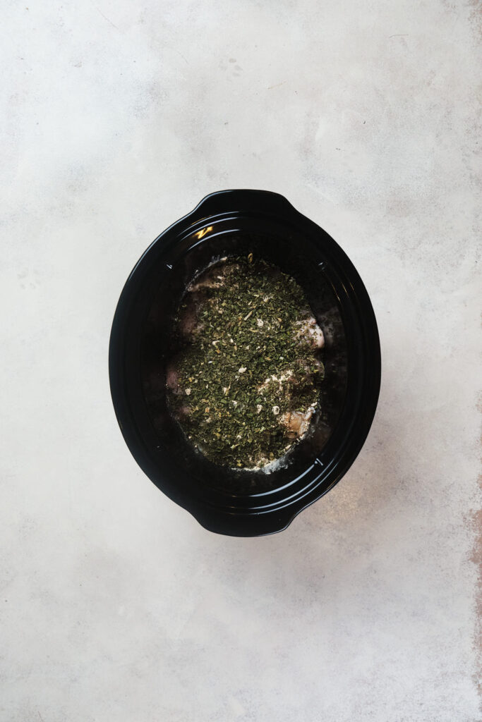 A black crock pot with chicken and herbs on a light grey surface.