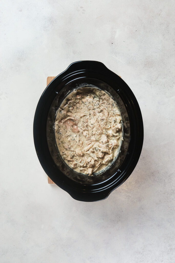 A creamy chicken mixture in a black crock pot on a light grey surface.