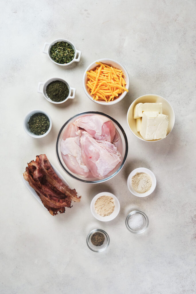 Assorted cooking ingredients including raw chicken, bacon, cheese, herbs, and spices arranged neatly in bowls on a light surface.