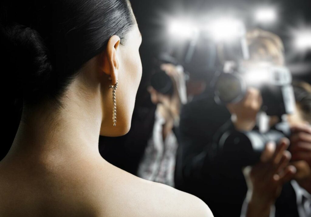 Woman on a red carpet event, photographed from behind, focusing on her earring, with photographers and camera flashes in the background.