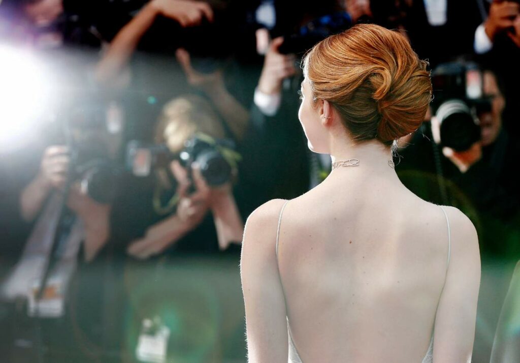 Celebrity with an elegant updo hairstyle facing away from the camera towards a crowd of photographers at a formal event.