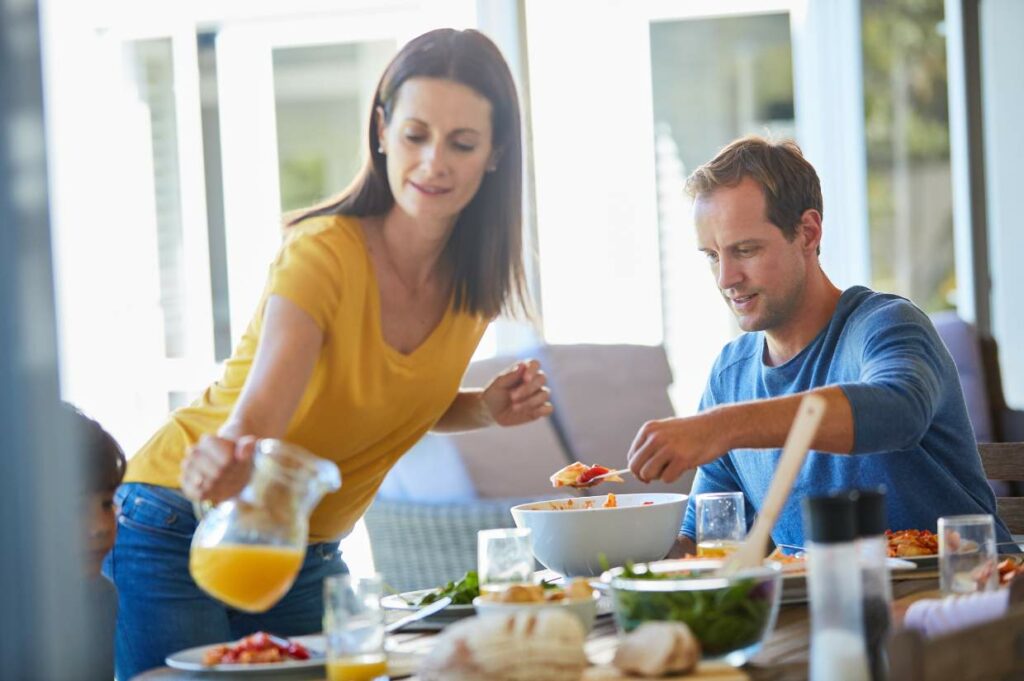 A woman pours juice while a man serves salad at a breakfast on a sunny patio.