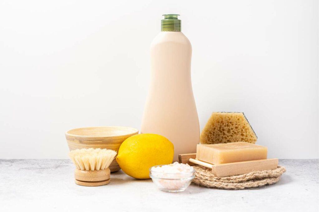 Assorted bath accessories including a bottle of liquid soap, bar soap, a brush, a wooden bowl, a sponge, a lemon, and bath salts, arranged on a light surface.