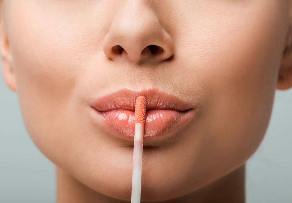 Close-up of a woman applying lip gloss with a brush on her lips against a gray background.