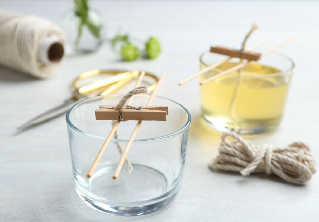 Two glass containers with wick-centering devices set up, one with wax and the other empty. Nearby are a pair of scissors, a roll of twine, and some loose string.