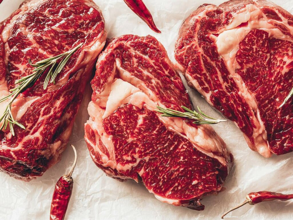 Three raw marbled steaks garnished with sprigs of rosemary and chili peppers, lying on parchment paper.