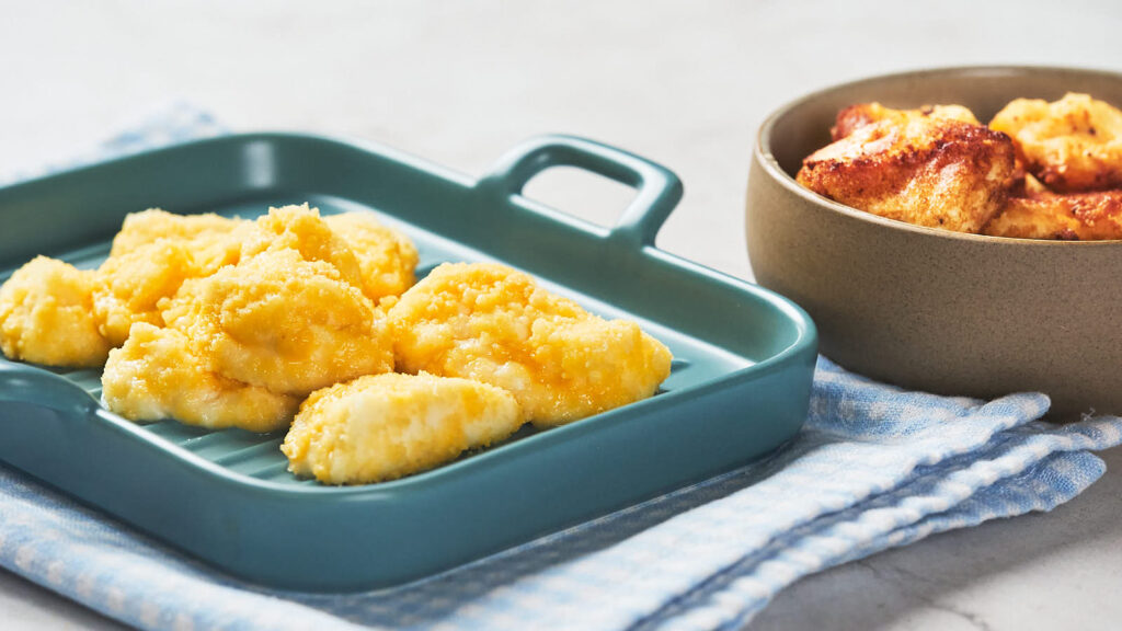 Two dishes of food are displayed: one has small portions of fried food on a blue tray, the other contains baked pieces of food in a brown bowl.