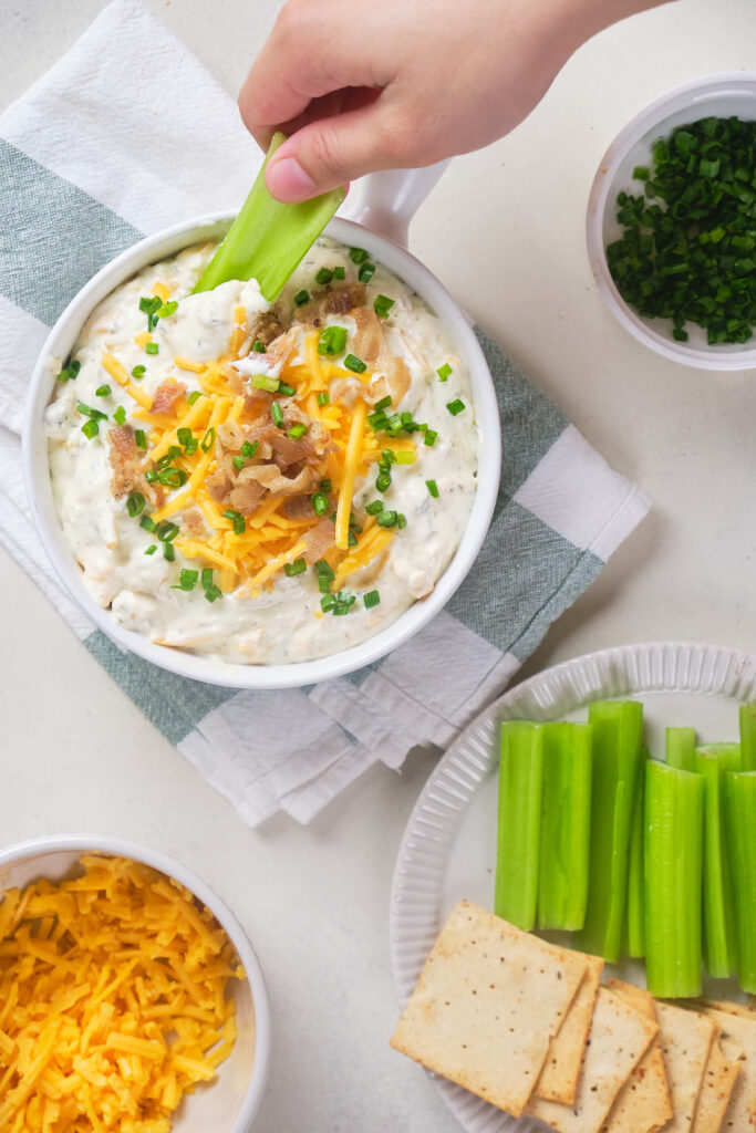 A hand dipping a celery stick into a bowl of creamy dip topped with shredded cheese and bacon.