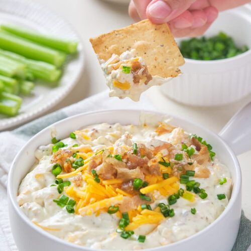 A hand holding a cracker dipped in a white bowl of creamy dip topped with cheese and bacon.