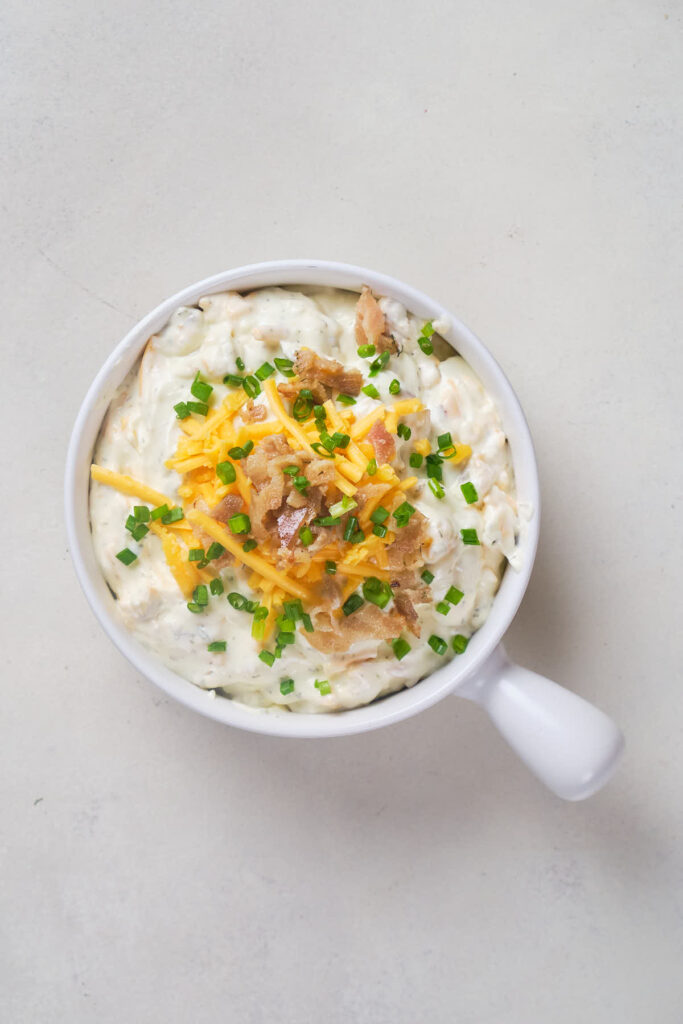 A white bowl filled with crack dip with shredded cheese and bacon bits on a light surface.