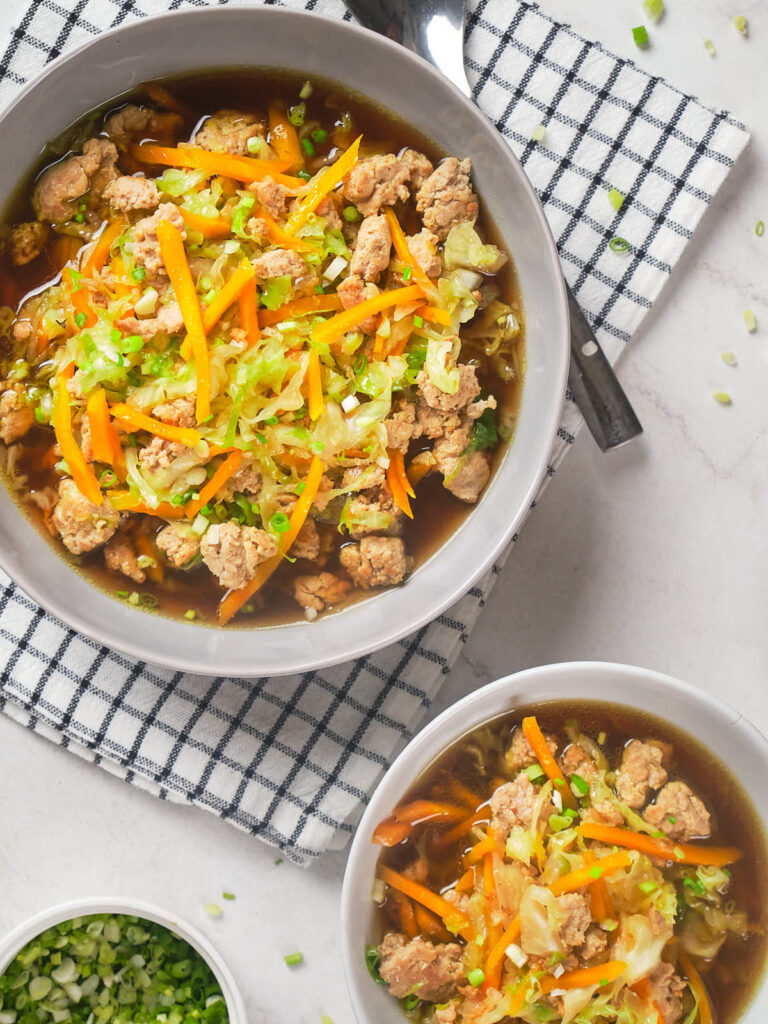 Two bowls of soup with ground meat, shredded carrots, and green onions, set on a checkered cloth with a spoon.