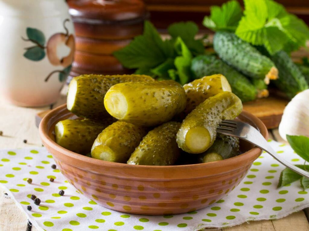 A bowl of pickles with a fork on a polka-dot cloth, surrounded by fresh cucumbers, black peppercorns, and greenery.