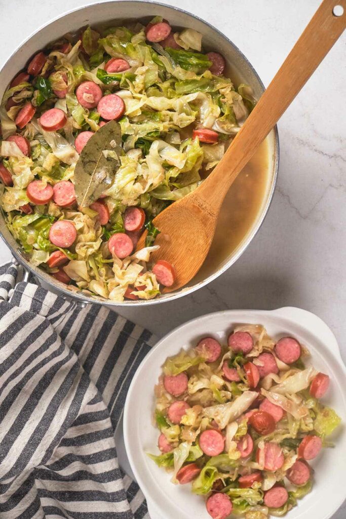 A pot and a bowl of cooked cabbage with sliced sausages, accompanied by a wooden spoon.