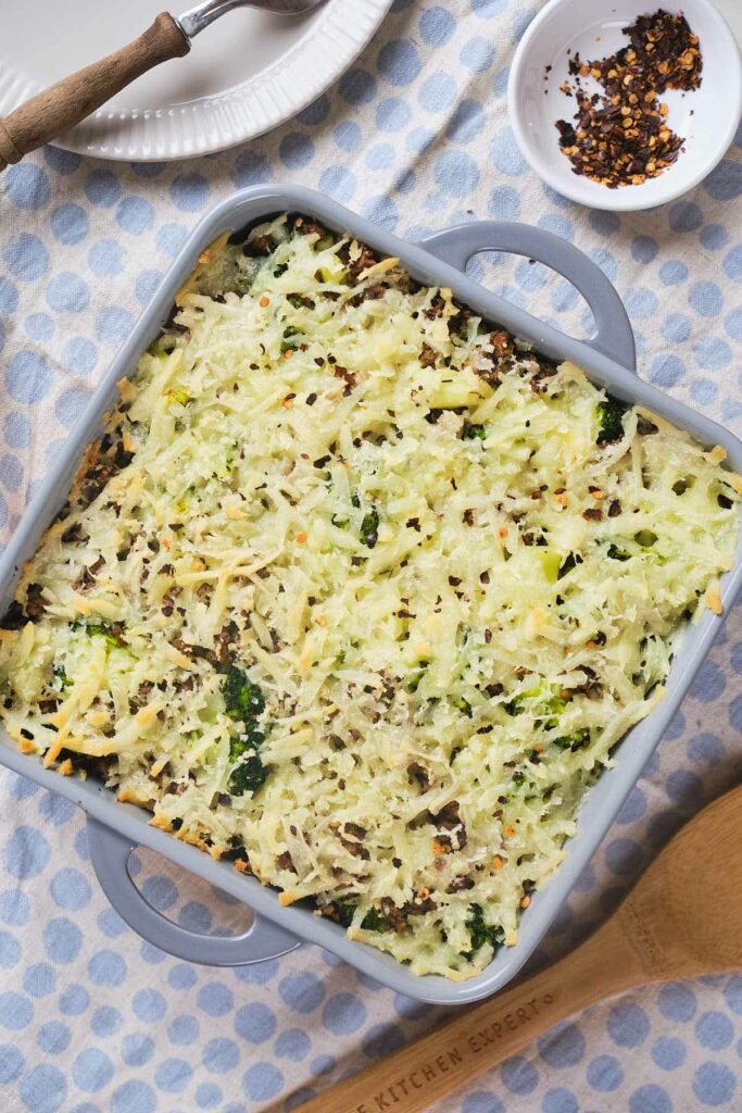 A gray baking dish contains a freshly baked casserole topped with shredded cheese and red pepper flakes, sitting on a blue and white polka dot cloth. A wooden spoon and a small dish are beside it.