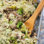 A casserole dish with a wooden spoon, featuring a mixture of cooked ground meat, broccoli, and melted cheese. A small bowl of red pepper flakes is in the background.