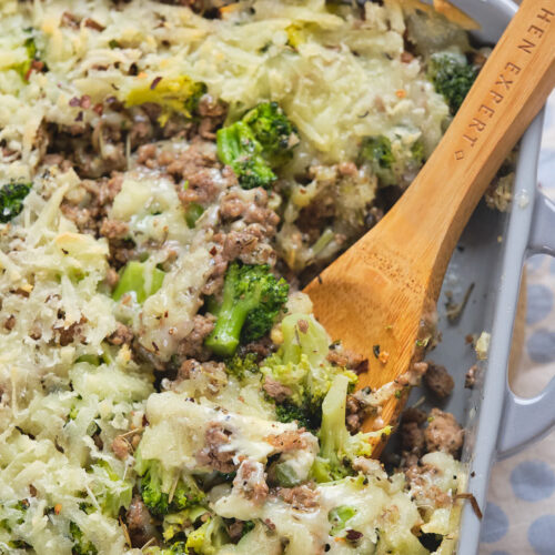 A casserole dish with a wooden spoon, featuring a mixture of cooked ground meat, broccoli, and melted cheese. A small bowl of red pepper flakes is in the background.